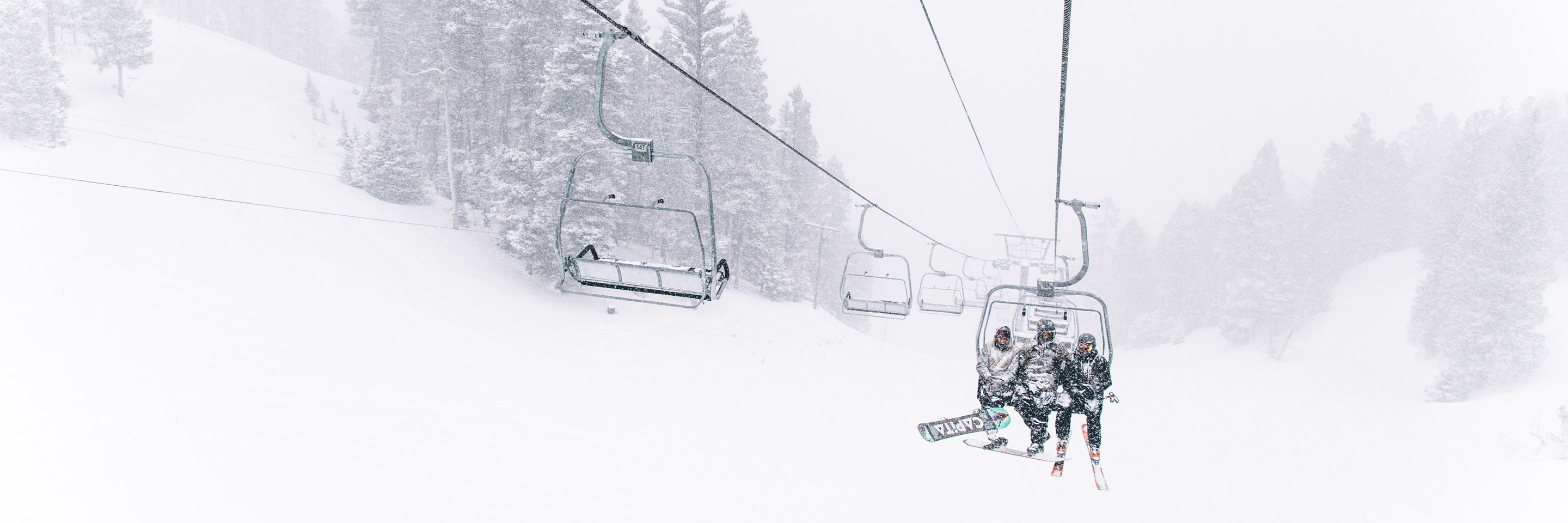 three skiers and snowboarders riding a chairlift in snow